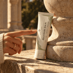 A hand points toward a tube of anceitn therapy recovery balm standing on a stone ledge outdoors. The tube is white with pale green abstract shapes and black text reading 'ANCIENT HERBS RECOVERY BALM'