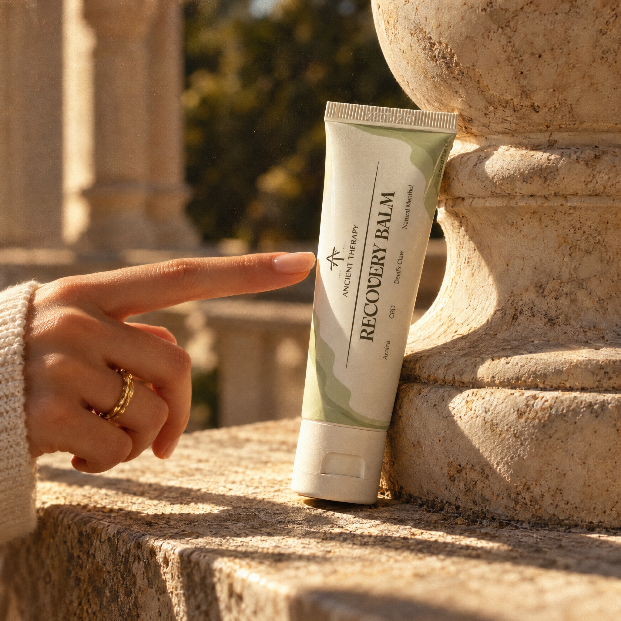 A hand points toward a tube of anceitn therapy recovery balm standing on a stone ledge outdoors. The tube is white with pale green abstract shapes and black text reading 'ANCIENT HERBS RECOVERY BALM'