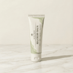 A tube of Ancient Therapy Recovery Balm standing upright on a light marble surface against a warm off-white background.
